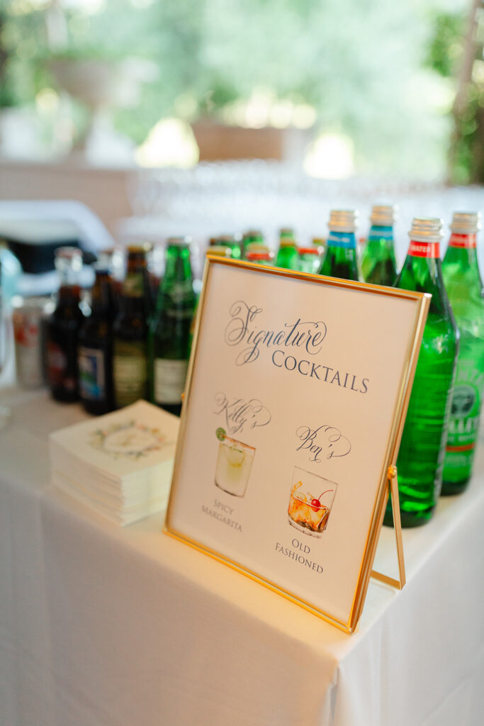 Signature cocktail sign beside bottles at the wedding bar.