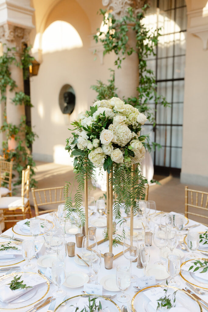Tall white floral centerpiece with greenery in a gold stand.
