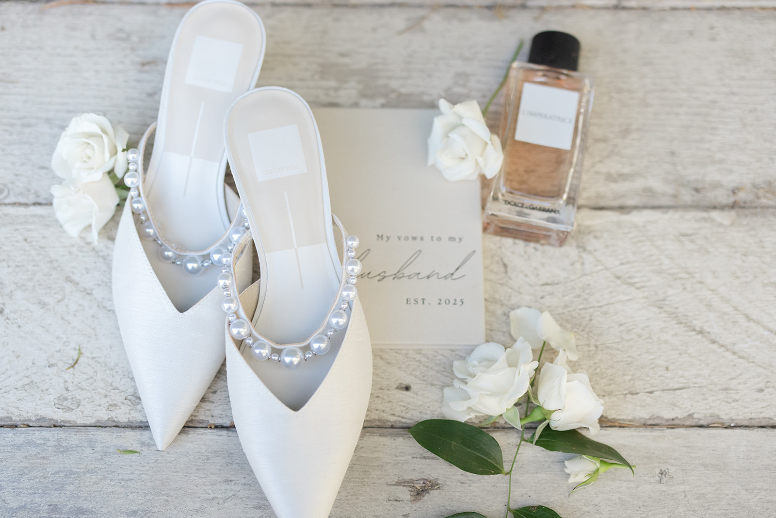 Pearl-trimmed bridal shoes, perfume, and white roses on a wooden table.