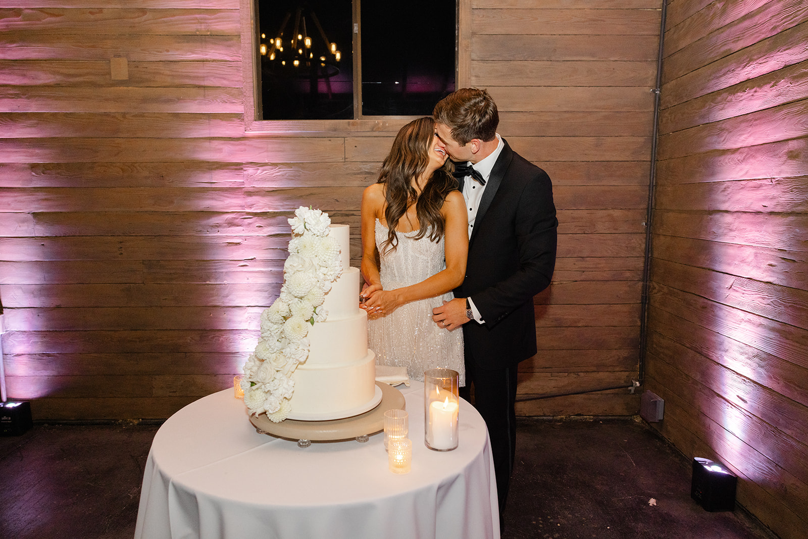 Jake kissing Zoe beside their white tiered wedding cake.