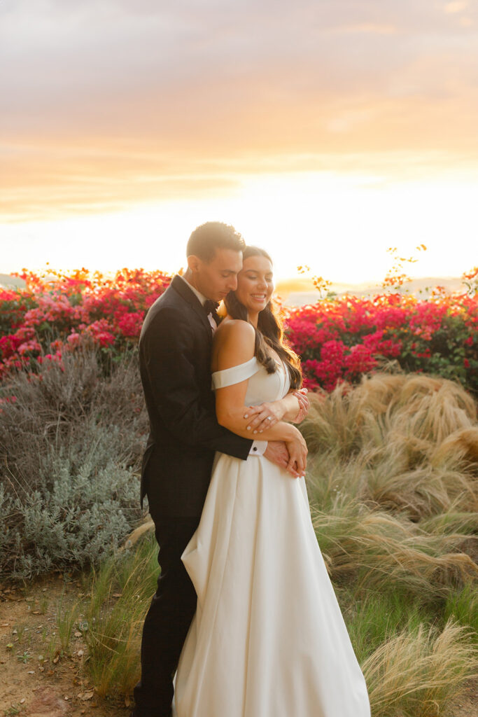 Samantha and Andrew embracing at sunset beside pink flowers at Quail Ranch.