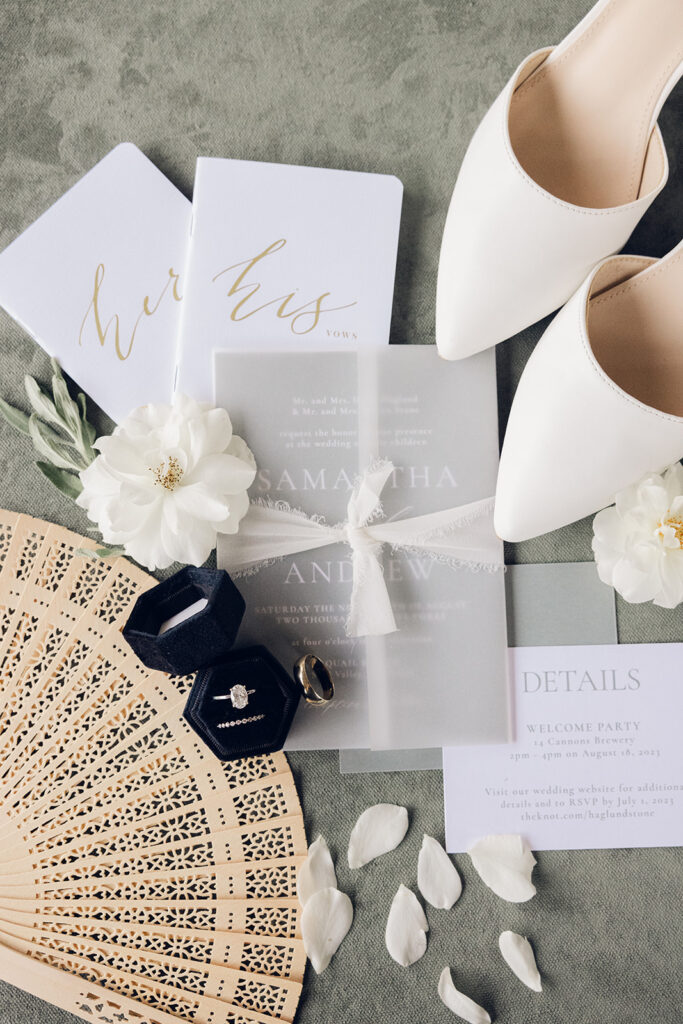 Samantha and Andrew’s wedding invitations, vow books, rings, white shoes, and wooden fan.