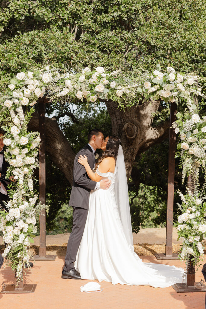 Samantha and Andrew kissing beneath the white-flower wedding arbor.
