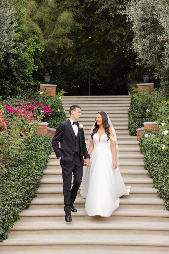 Samantha and Andrew walking hand in hand down the flower-lined garden staircase.
