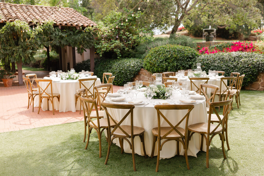 Outdoor reception tables with white linens, flowers, and wooden cross-back chairs.