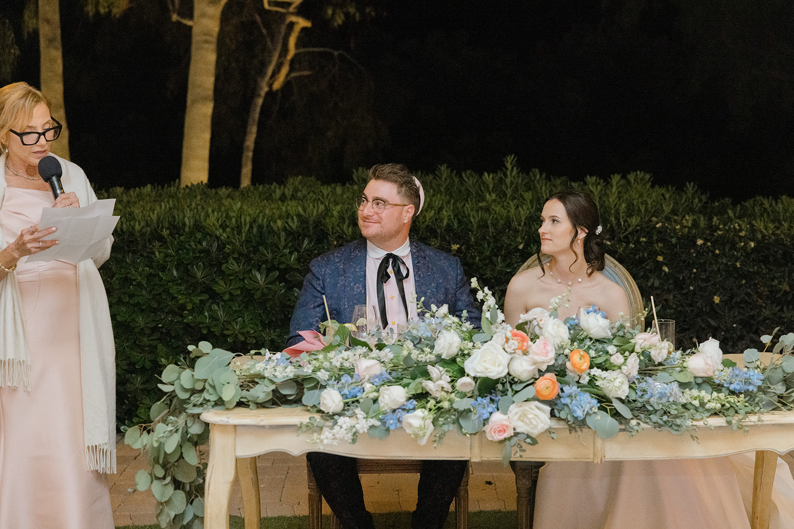Jordan and Sara listening to a toast at their flower-covered sweetheart table